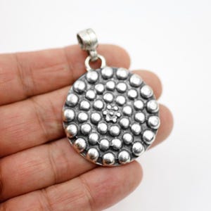 Peut inclure: Un pendentif circulaire de couleur argentée présentant une surface texturée couverte de nombreuses petites sphères en relief disposées en cercles concentriques rayonnant autour d'un groupe central de sept points, surmonté d'une bélière.