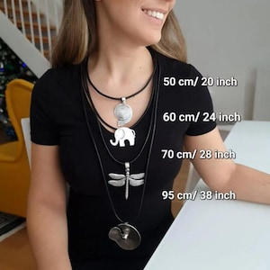 Puede incluir: Una colección de cuatro collares en capas con colgantes plateados. Los colgantes incluyen una espiral, un elefante, una libélula y un diseño circular. Los collares miden 50, 60, 70 y 95 cm.