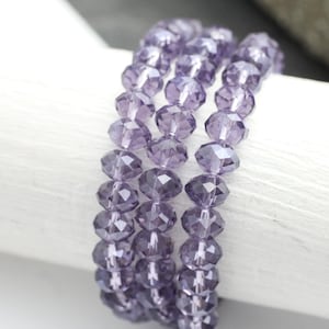 May include: Close-up of several strands of lavender-coloured faceted beads. The beads are strung together, forming bracelets. The beads have a sparkling, crystal-like appearance. The bracelets are displayed on a white surface.