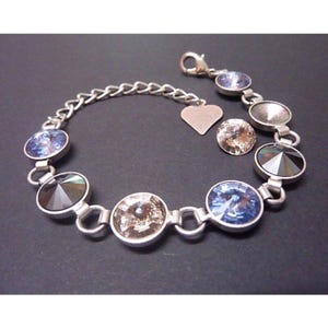 May include: A silver-toned bracelet featuring a series of circular, faceted crystals in shades of blue, grey, and champagne. The bracelet includes a delicate chain, a lobster claw clasp, and a small, smooth heart-shaped charm attached to the links.