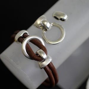 May include: A brown leather bracelet with silver-toned metal accents. The bracelet features a unique knot design with a circular clasp and two additional silver pieces. The leather appears to be a rich, warm brown color.