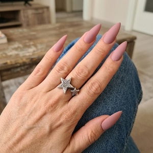 Könnte beinhalten: Ein silberfarbener Ring mit zwei sternförmigen Elementen, wobei der größere Stern eine texturierte, gepunktete Oberfläche aufweist. Der Ring wird an einem Finger mit langen, mandelförmigen Nägeln in mattem, gedecktem Mauve-Rosa getragen.