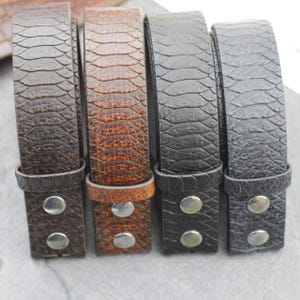 May include: Four belts with a textured snakeskin pattern in shades of dark brown, medium brown, charcoal gray, and light gray. Each belt features a matching loop and two silver-toned metal snap buttons at the end for secure fastening.