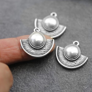 Pode incluir: Três pingentes de metal prateado. Cada pingente apresenta uma forma de semicírculo com uma superfície texturizada e um elemento circular no centro. Uma pequena argola na parte superior para prender a uma corrente ou outro componente de joalheria.