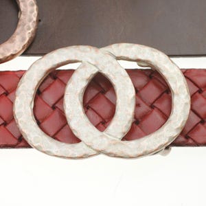 May include: A close-up of a red woven leather belt with a silver-colored double-circle buckle. The buckle has a hammered texture. The belt is a deep red color, and the buckle is a light silver color.