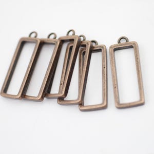 May include: Several rectangular bronze-colored metal pendant blanks with a loop at the top for jewelry making. The open frames are empty, ready for embellishment with resin, beads, or other decorative elements. Ideal for DIY projects.