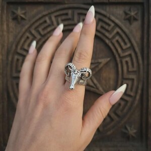 May include: A silver-toned metal ring featuring a detailed ram head design with curved horns. The ring has a textured band and a polished finish on the ram's face, showcasing intricate craftsmanship and a bold, sculptural aesthetic on the finger.