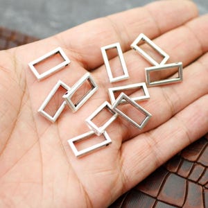Puede incluir: Una colección de pequeños dijes rectangulares de metal plateado. Cada dije tiene un centro hueco y abierto. Los dijes están dispersos sobre una superficie de tono claro, lo que sugiere que son suministros para manualidades o joyería.