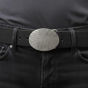 Könnte beinhalten: Ein schwarzer Gürtel mit strukturierter Oberfläche und einer großen, ovalen Gürtelschnalle aus silberfarbenem Metall. Die Schnalle ist zweigeteilt mit einem gitterartigen Rautenmuster auf der linken Seite und vertikalen Linien rechts.