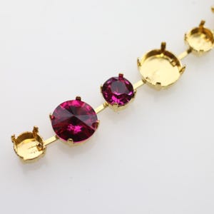 May include: A gold-toned metal chain featuring a series of circular settings. Two of the settings are filled with round, faceted, deep magenta-colored stones, while the remaining settings are empty, showing the gold prongs and base.