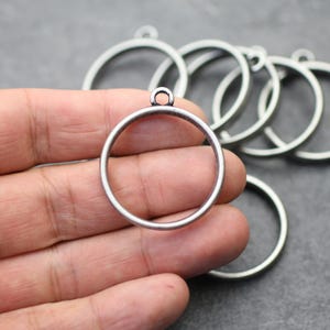 以下が含まれることがあります： ジュエリー作りに使用する、上部に小さなループが付いたシルバーカラーの金属製円形ペンダント。 ペンダントは丸く、シンプルでミニマルなデザインです。 いくつかのペンダントが見え、一部は手で持たれています。