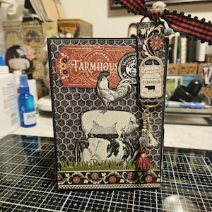 May include: A rectangular scrapbook album featuring a black and white honeycomb pattern background, a rooster and cow illustration, and a red label with the text Happiness is Home Farmhouse. A tag with a milk can and cow graphic hangs from a red plaid ribbon.