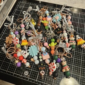 Op de afbeelding: Een verzameling kralen-sleutelhangers met diverse bedels zoals spoken, pompoenen, cactussen, bloemen, snoep en teddyberen. Elke sleutelhanger is versierd met zilveren lintstrikjes en roségouden metalen ringen, met kleurrijke siliconen en kunststof kralen in diverse vormen.