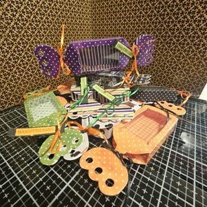 Könnte beinhalten: Eine Sammlung von bonbonförmigen Geschenkboxen aus Papier in Lila, Grün und Orange mit Punkten und Sichtfenstern. Jede Schachtel ist mit einem orangefarbenen oder grünen Band versehen und trägt ein grünes Etikett mit der Aufschrift Happy Halloween.