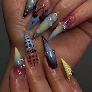 May include: A hand showing long, pointed acrylic nails with diverse designs. The nails feature gradients, polka dots, cherries, and silver embellishments. Colours include blue, yellow, pink, and burgundy.