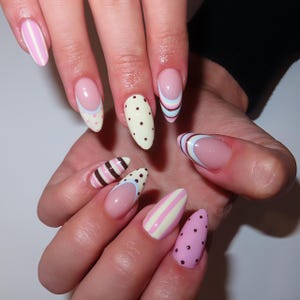 May include: A hand displaying almond-shaped nails with various designs. The nails feature stripes, polka dots, and French tips in pastel shades of pink, white, brown, and blue. The nails are well-manicured and have a glossy finish.