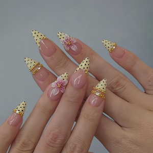 Peut inclure: Ongles artificiels en forme d'amande. Les pointes sont peintes avec une base jaune et des pois noirs. Certains ongles présentent des fleurs 3D roses et blanches. Des bijoux dorés sont ajoutés à certains ongles.