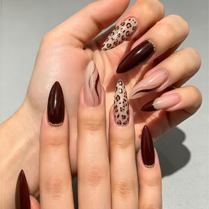 May include: Almond-shaped nails featuring a mix of glossy dark brown polish, leopard print patterns on a nude base, and nude nails with thin brown swirl accents. Several nails are accented with small gold beads at the cuticle line.