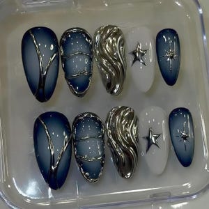 Op de afbeelding: Een set van tien kunstnagels in een doorzichtige plastic container. De nagels hebben verschillende ontwerpen, waaronder een blauw-zilveren gradiënt, zilveren golfpatronen, wit met een zilveren ster en een blauwe gradiënt met een zilveren ster.