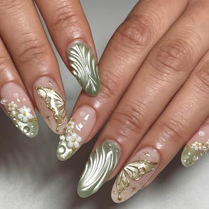 May include: Almond-shaped nails featuring a nude base with metallic sage green 3D wave patterns, gold butterfly wing decals, small white floral accents, and clear raised droplets. The design incorporates tiny gold star motifs and pearl-like beads.