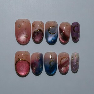 Puede incluir: Un conjunto de diez uñas artificiales con temática celestial. Las uñas presentan un degradado de tonos rosa, morado y azul, adornadas con medias lunas doradas, estrellas y detalles brillantes. Las uñas son de forma ovalada.