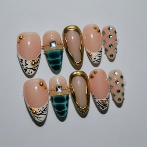 May include: A set of ten almond-shaped artificial nails. The nails feature a variety of designs, including gold accents, zebra print, teal patterns, and clear gems. Some nails have a nude base with gold studs.