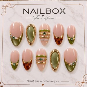 May include: A set of ten almond-shaped press-on nails on a white display card labeled NAILBOX For You. The nails feature a nude base with olive green tips, gold metallic accents, small green rhinestones, white pearls, and brown tortoiseshell patterns.