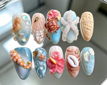 Puede incluir: Un juego de diez uñas postizas con diseños 3D de temática oceánica. Las uñas muestran motivos que incluyen un caballito de mar, conchas, una tortuga, una estrella de mar, un tentáculo de pulpo, una flor de hibisco y detalles iridiscentes en azul, rosa y beige.