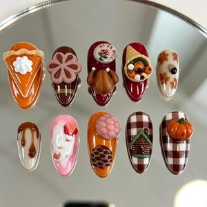 May include: A set of ten press-on nails featuring 3D embellishments including a pumpkin pie slice, a brown flower, a roasted turkey, a cornucopia, autumn leaves, caramel drizzle, a strawberry cream design, berries, a cabin, and a pumpkin.