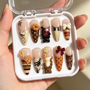 May include: A set of ten almond-shaped press-on nails in a white plastic case. The designs feature autumn-inspired motifs including brown and cream stripes, polka dots, floral swirls, musical notes, 3D bows, leaves, and small animal figures.