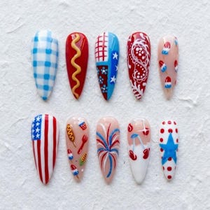 May include: A set of ten long, almond-shaped artificial nails with various patriotic designs. The designs include gingham, hot dogs, fireworks, the American flag, and stars. Colors include red, white, blue, and tan.