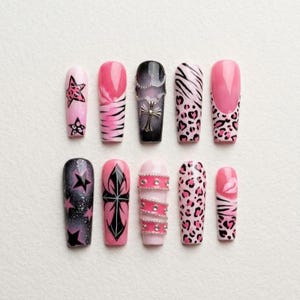 May include: A collection of ten long, coffin-shaped false nails. Designs include pink and black star and cross patterns, zebra stripes, leopard print, and a black nail with a silver cross pendant. Some nails have rhinestones.