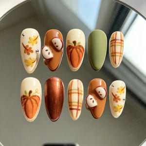 May include: A set of ten press-on nails featuring autumn-themed designs including cream backgrounds with yellow and orange leaves, solid metallic copper, solid olive green, brown with 3D apple slices, orange pumpkins, and a cream, brown, and green plaid pattern.