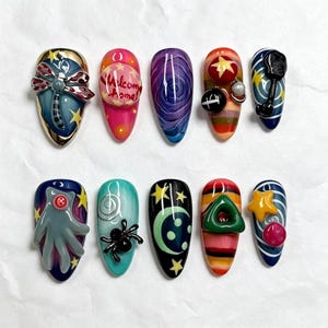 May include: A set of ten almond-shaped press-on nails featuring diverse 3D designs, including a dragonfly, a pink sphere with the text Welcome home, a purple swirl, a black key, a gray hand with a red button, a black spider, and stars.