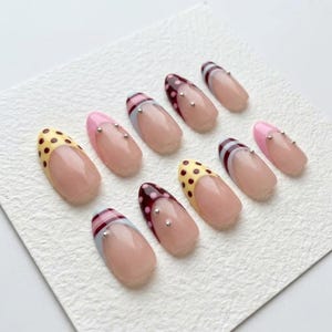 Op de afbeelding: Een verzameling van tien kunstnagels met diverse ontwerpen. Sommige nagels hebben een nude basis met kleurrijke tips, waaronder gele stippen, roze en bordeaux met zilveren accenten. Andere hebben gestreepte patronen.
