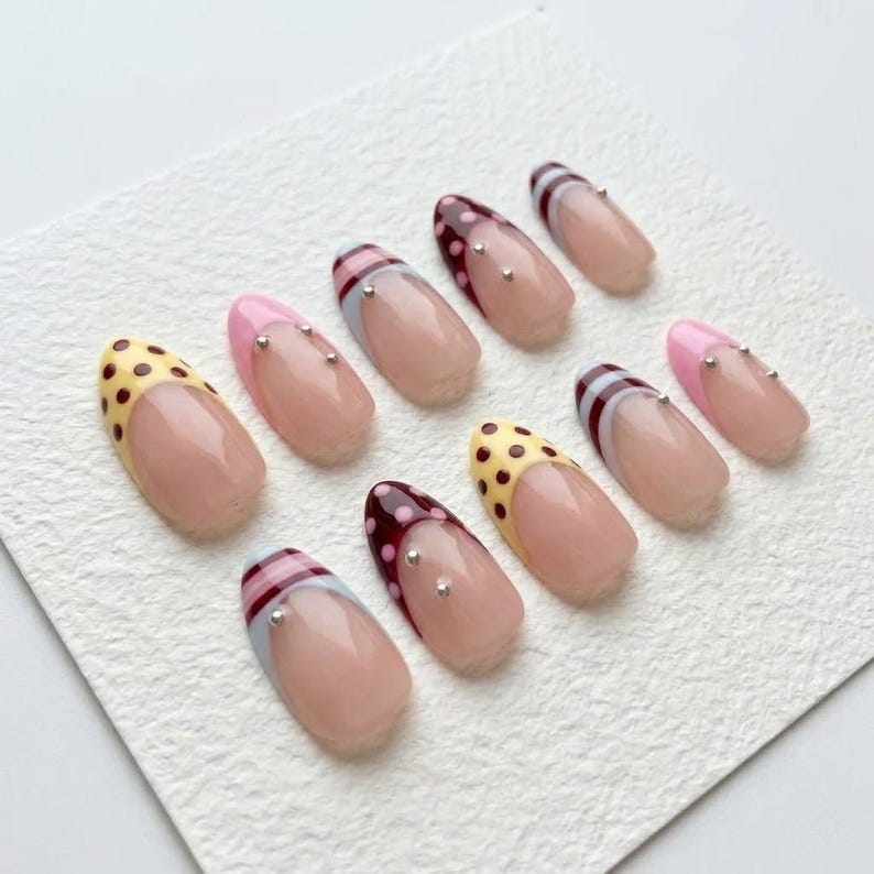 Op de afbeelding: Een verzameling van tien kunstnagels met diverse ontwerpen. Sommige nagels hebben een nude basis met kleurrijke tips, waaronder gele stippen, roze en bordeaux met zilveren accenten. Andere hebben gestreepte patronen.