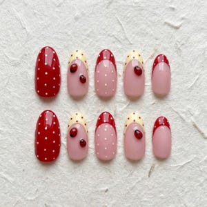 May include: A set of ten artificial nails in various designs. Two nails are solid red with white polka dots. Other nails feature a pink base with white polka dots, a red tip, and a small ladybug accent. The nails have an oval shape.