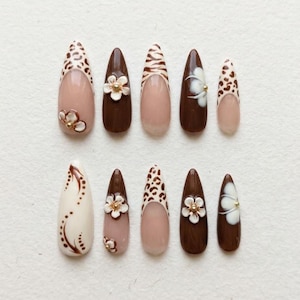 May include: A collection of ten almond-shaped false nails. The nails feature various designs, including leopard print, floral accents, and solid colors in shades of brown, beige, and white. Some nails have a nude base with detailed patterns.