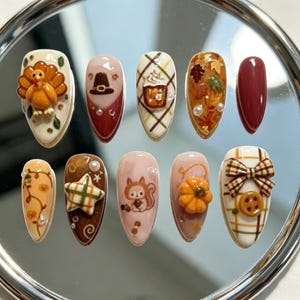 May include: A set of ten almond-shaped press-on nails featuring autumn-themed designs including a 3D turkey, a pilgrim hat, plaid patterns, fall leaves, a squirrel, a pumpkin, a brown bow, and a button, accented with small white pearls.