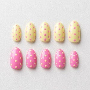 May include: A set of ten oval-shaped false nails. Five are yellow with pink polka dots, and five are pink with yellow polka dots. The nails are arranged in two rows of five.