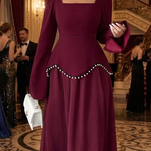 Puede incluir: Un vestido de noche burdeos con escote cuadrado y mangas largas. La cintura está adornada con un ribete de cuentas y la falda fluye elegantemente. Se sostiene un bolso blanco. El escenario es un gran salón de baile.