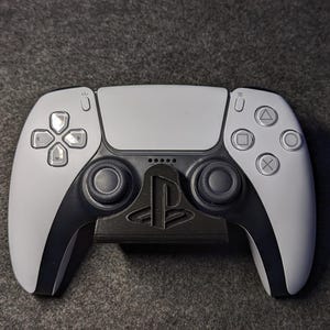 May include: A gray and black PlayStation 5 controller resting on a black stand. The controller features a white faceplate with black accents and the PlayStation logo on the stand.