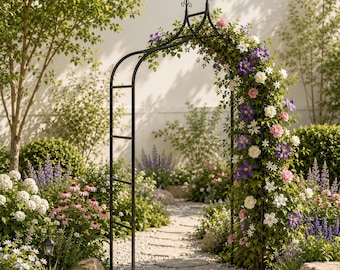 Op de afbeelding: Een zwarte metalen tuinboog met een puntige top, begroeid met een klimplant vol paarse, witte en roze bloemen. De boog staat over een stenen pad in een tuin met groen gebladerte en diverse bloeiende planten.
