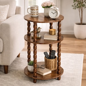 May include: A three-tiered, round, wooden side table with a dark brown finish. The table features turned legs and holds decorative items like a mug, alarm clock, flowers, a bird figurine, books, a candle, a small plant, a basket with remotes, and a pair of glasses.