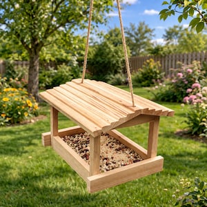 May include: A wooden bird feeder with a sloped roof and a rectangular feeding tray. The feeder is suspended by rope and filled with birdseed. The natural wood color blends with the outdoor setting of green grass and trees.