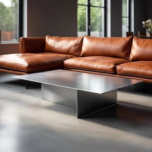 Puede incluir: Un salón moderno con un gran sofá seccional de cuero marrón y una mesa de centro rectangular de metal plateado. La habitación tiene grandes ventanales y un suelo de hormigón pulido, creando una estética minimalista y contemporánea.