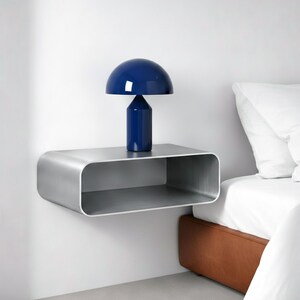 Peut inclure: Scène de chambre moderne avec une table de chevet flottante en métal brossé aux bords arrondis. Une lampe bleu foncé en forme de champignon est posée sur la table. Un lit blanc et une tête de lit marron sont visibles en arrière-plan.