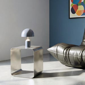 May include: A modern, brushed metal side table with a rounded square design. A small, light blue mushroom-shaped lamp sits on top. The lamp has a cylindrical base and a white light. The background features a blue wall and a black leather chair.