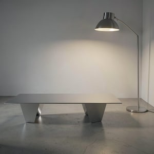 May include: A modern, brushed metal coffee table with a rectangular top and geometric legs. A silver-toned floor lamp with a domed shade is positioned to the right, casting light on the table. The room has a minimalist design.