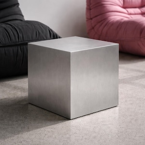 Peut inclure: Une table d'appoint en forme de cube en métal brossé. La table est de couleur gris argenté uni et repose sur un sol en béton. L'arrière-plan comprend un fauteuil noir et un fauteuil rose.